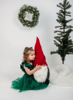 Toddler poses with gnome near christmas tree for holiday photo