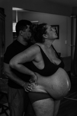 Photo captures dad supporting mom as she labors during childbirth at a home birth; Man applies counterpressure to womans back during a contraction