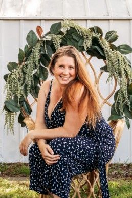 Woman posed sitting in chair surrounded by flowers smiles at the camera