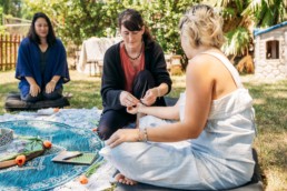 Three women gather in a circle format. One woman ties a cord around another's wrist. There are flowers and journals set in front of the women