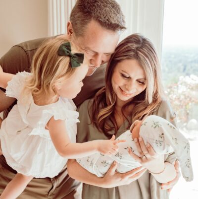 Dad holds toddler while mom holds newborn. All family members gaze down and smile at newborn baby