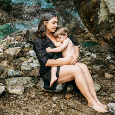 Mother sits at foot of a waterfall and breastfeeds toddler while holding him close. She has her eyes closed and looks peaceful