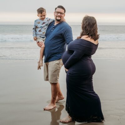 Pregnant mom, dad, and older child walk along the beach shoreline. Dad and mom gaze at each other while mom holds her pregnant belly. Older child looks downward.