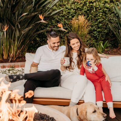Family of three sit on a white couch outside while a dog is at their feet. They are in front of a fire pit. Mom and daughter drink from mugs. Dad and mom look at daughter.
