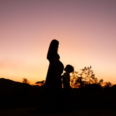 Silhouette of mom with pregnant belly and older child kissing her belly at sunset