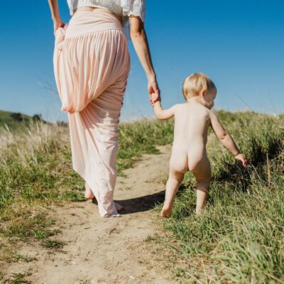 Image of toddler picking at grass while holding mother's hand. Mother is pulling up a long pink skirt as she walks