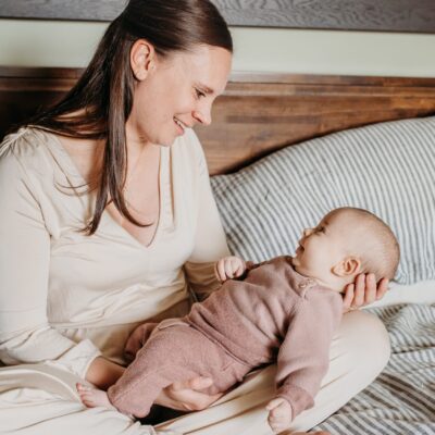 Image of mom and baby sat on the bed. Baby is in mom's lap and they stare into each other's eyes. The image is brightly lit and baby is smiling.