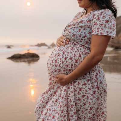 Pregnant woman holds her belly with sun setting behind her. She stands on a shoreline gazing out to the water.