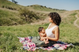 Mother has a peaceful look on her face as she sits on a blanket amongst rolling green hills and breastfeeds her baby