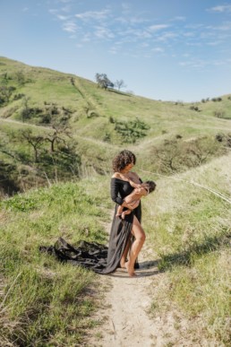 Mother in long black gown hold baby while breastfeeding on a trail amongst rolling green hills