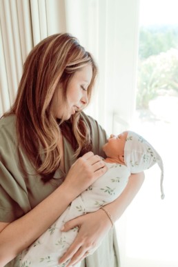 Mother holds newborn baby who is wrapped in swaddle near the window. Mom and baby gaze into each others eyes