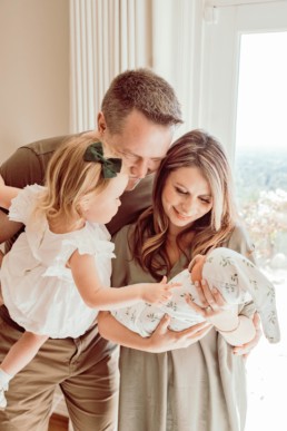Dad holds toddler while mom holds newborn. All family members gaze down and smile at newborn baby