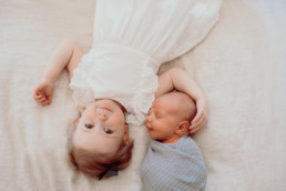 Newborn and toddler lay opposite directions. Newborn is wrapped and looking at sister. Photo taken from above children.