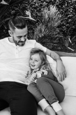 Daughter snuggles into her father's side while smiling and holding her stuffed bunny, dad looks down at daugher