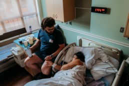 Image of laboring women closing eyes while laying on hospital bed. Father is touching her hand and gazing down at mom.