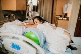 Image of laboring women drapped over peanut ball on top of hospital bed with eyes closed.
