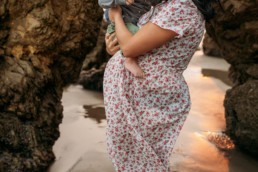 Pregnant women holds toddler over pregnant belly in front of rocky beach.