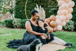 Mother and daughter sit snuggled up to each other, faces turned toward one another while smiling. Their dog sits out of focus in front of them.