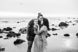 Dad, mom, and baby are standing on the beach. Mom is holding baby's face and dad is kissing mom on the cheek