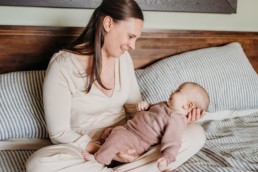 Image of mom and baby sat on the bed. Baby is in mom's lap and they stare into each other's eyes. The image is brightly lit and baby is smiling.