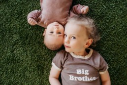 Image of child laying opposite of a baby with their heads touching. The child is wearing a shirt that says 