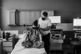 Laboring women sits cross legged on a hospital bed with head dipped down. Father is standing beside her with hands on her back for support. Both are wearing black masks.