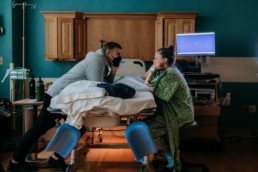 Laboring woman sits on a stool while leaning over a hospital bed. Father leans over the opposite side of the bed looking at woman. Woman has her head in her hands and eyes closed. Image of hospital birth equipment in the background.