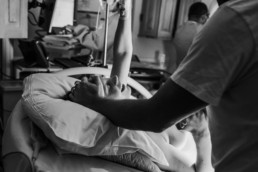 Black and white image of a mother laying on a hospital bed right after birth with her hand in the air in triumph. She has an expression of happy relief on her face. Dad is holding one of mom's hands and his other hand is on her head.