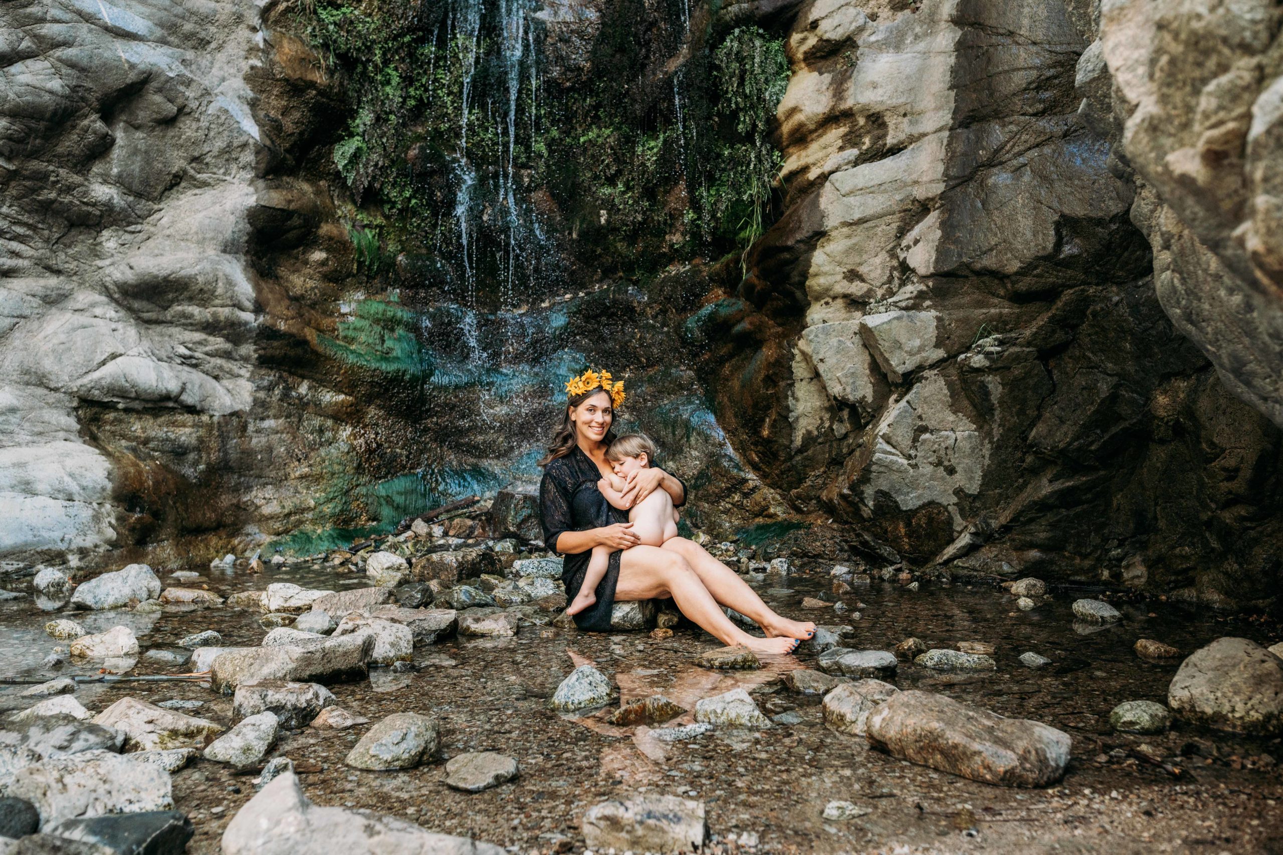 Mother sits at foot of a waterfall and breastfeeds toddler while holding him close. Mother is wearing a flower crown and smiling at the camera