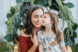 Mother smiles and closes eyes while her child kisses her on the cheek. They sit on a chair adorned with greenery