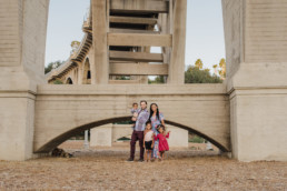 Image of family of five in front of a large bridge. Two older children are standing, leaning against mom while dad holds the youngest. All are looking at the camera and smiling.