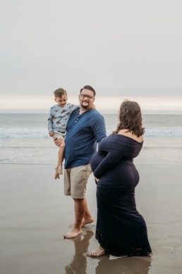 Pregnant mom, dad, and older child walk along the beach shoreline. Dad and mom gaze at each other while mom holds her pregnant belly. Older child looks downward.