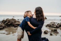 Pregnant mom hold her belly with one hand and her older child with another hand as they stand on a beach shore. Mom and son gaze into each others eyes.