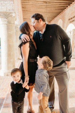 Family of four stand. Mother and father are kissing. Older child is looking up at parents. Younger child is looking at camera