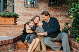 Family of hour are in a hug. Dad and older child look at each other. Younger child looks away while mom gazes at the camera. Family sits on brick steps