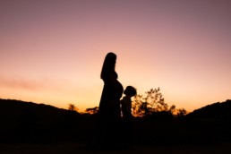 Silhouette of mom with pregnant belly and older child kissing her belly at sunset