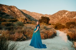 Pregnant woman stands in front of a mountainscape while holding her belly looking at the camera
