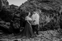 Pregnant woman stands looking at partner who is looking back at her. Both are resting hands on pregnant belly